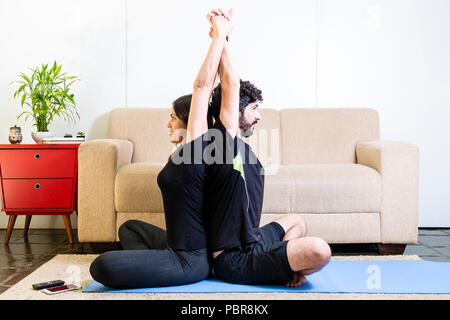 Beautiful hetero caucasian couple in black clothes on blue yogamat doing sit back-to-back pose with arms up Stock Photo