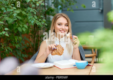 Smiling girl with blonde hair eating croissant, looking at camera. Thick book laying on wooden surface in front of her with blue coffee cup. Model wearing white blouse decorated with pearls, smiling. Stock Photo