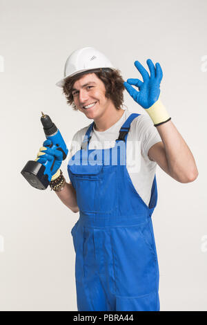 Studio portrait of happy builder laughing at camera with screwdriver wearing gloves and white helmet. Stock Photo