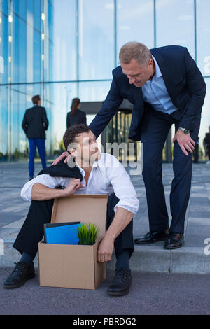 Fired business man sitting frustrated and upset on the street near office building with box of his belongings. He lost work. Other businessman comfort Stock Photo