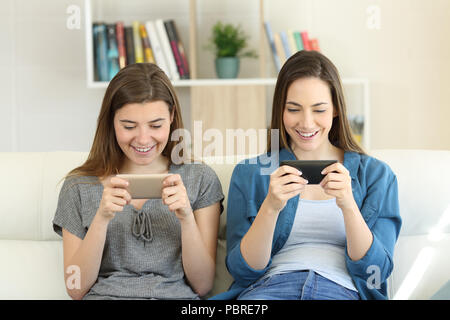 Two friends playing online games with smartphones sitting on a couch in the living room at home Stock Photo