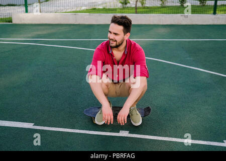 Smiling handsome fashionable guy dressed in a red shirt and shorts sitting on a skateboard on basketball court, looking away Stock Photo