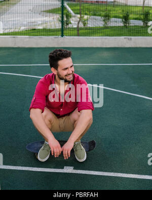 Smiling handsome fashionable guy dressed in a red shirt and shorts sitting on a skateboard on basketball court, looking away Stock Photo