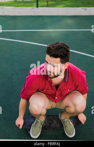 Smiling handsome fashionable guy dressed in a red shirt and shorts sitting on a skateboard on basketball court, looking away Stock Photo