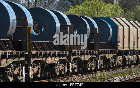 The back of a long and heavy rail freight train of wagons and flatbed trucks carrying large steel coils from a steelworks Stock Photo