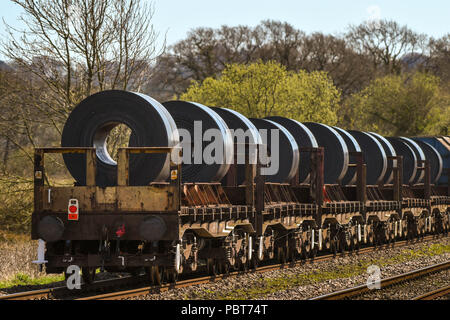 The back of a long and heavy rail freight train of flatbed trucks carrying large steel coils from a steelworks Stock Photo