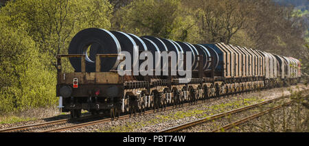 The back of a long and heavy rail freight train of wagons and flatbed trucks carrying large steel coils from a steelworks Stock Photo