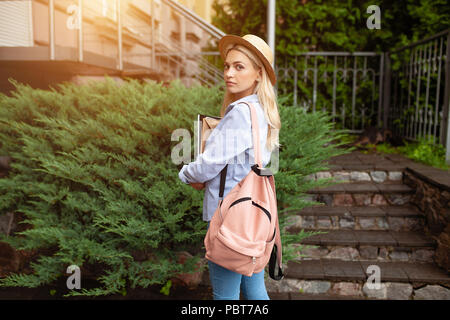 A beautiful student blonde in a straw hat with a pink backpack behind her back and books in her hands is standing in front of the university entrance  Stock Photo