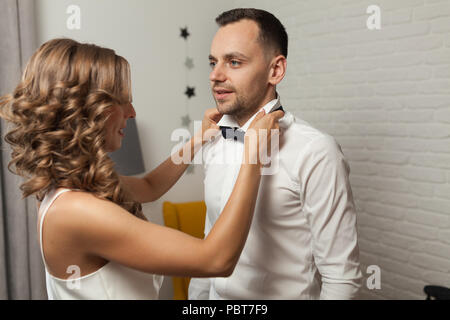 the bride and groom in the morning at the hotel on a wedding day. Dressing a newly-married couple Woman help fixing bowtie to groom. Stock Photo