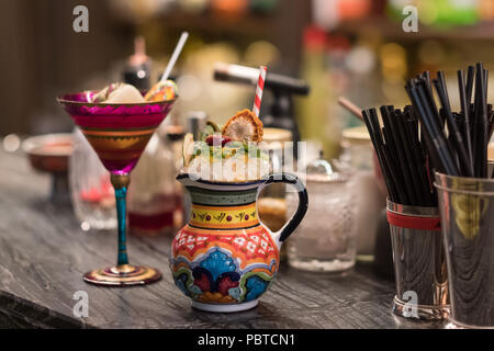 Fancy cocktails in red patterned wineglass and original painted jar are standing on bar counter with cocktail tools and ingredients. Stock Photo