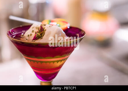 Alcoholic cocktail in red patterned glass with ice cube, dry rose flowers and candy on bar counter. Sweet fancy low alcohol drink. Stock Photo