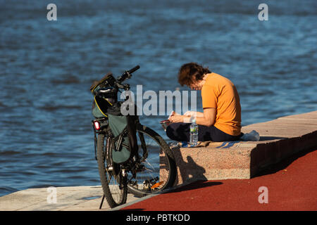Moscow. Unidentified woman enjoys reading a book by the Moscow-river on a sunny summer day. Her bicycle stands nearby. Stock Photo
