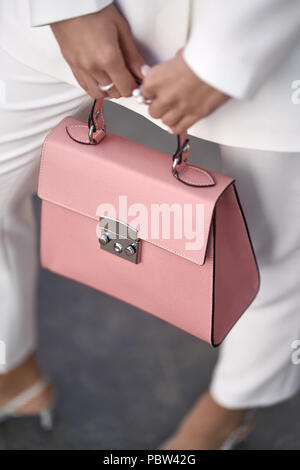 Nice girl is posing with a coral bag on the wall background outdoors. She wears a white pantsuit with light shoes. Closeup vertical photo. Stock Photo