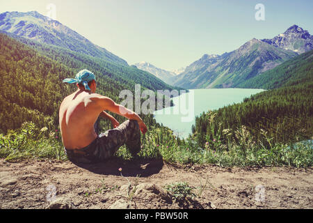 Calm man sitting on a rock on vacation looking at the lake and mountains on a summer evening. Mountain lake Kucherlinskoe from above, Altay, Russia. Stock Photo