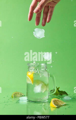 A woman's hand throws a piece of ice into a glass jar with a natural lemonade. Ice cubes fall into a glass jar with a splash and drops in different directions. Stock Photo