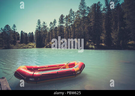 Inflatable boat on water on the river or lake near the shore in a clear summer day, the bright reflections on the waves. Red boat at the wooden bridge Stock Photo