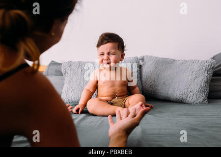 Annoyed baby crying sitting on the bed in the room wearing shorts, mom is in the frame of the image animating him, frustration concept Stock Photo