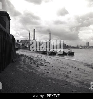 1950s, historical, the tide is out as barges sit on the south-bank of the river Thames by a small pebbly beach with a view  from the east of a working Deptford Power Station in the distance, London, England, UK. Designed by Sebastian de Ferranti, Deptford is know as the world's first central power station. Coal fired, it was unprecedented in both its scale and high voltage and it continued operating until the late 1960s. Stock Photo