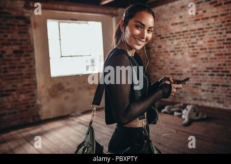 Portrait of a young woman in black sportswear listening to music from smart phone in hands and carrying a sports bag. fitness woman at gym looking at Stock Photo