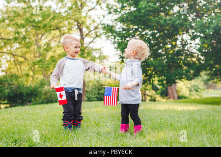 Happy adorable little blond Caucasian girl and boy smiling laughing holding hands and waving American and Canadian flags, outside in park, celebrating Stock Photo