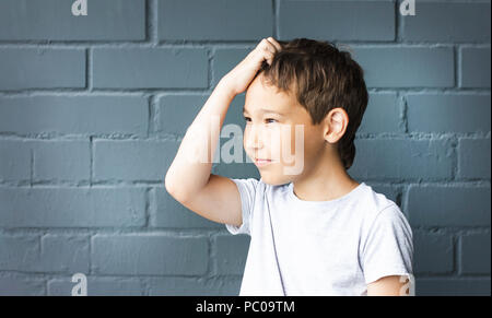 Boy 8 years old with freckles looking into the distance, thinking and scratching his head against the gray brick wall Stock Photo