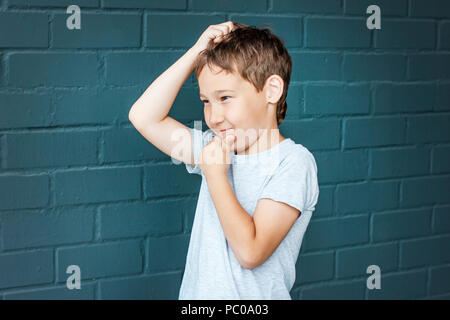 Boy 8 years old with freckles looking into the distance, thinking and scratching his head against the gray brick wall Stock Photo