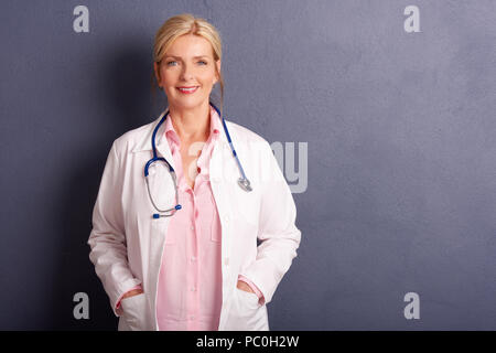 Shot of smiling female doctor wearing medical gown while standing at grey background with copy space. Stock Photo