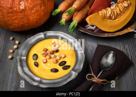 Tasty pumpkin and carrot cream soup served with croutons and pumpkin seeds in metal pewter bowl. Ingredients and toppings around the bown on gray rust Stock Photo