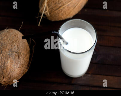 A glass of coconut milk with black straw and two whole coconuts on wooden background. Stock Photo