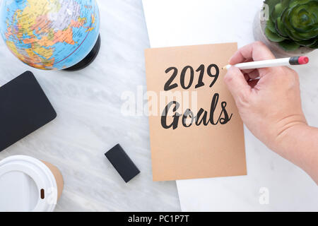 Hand holding pen writing 2019 Goals on brown paper with blue globe,blackboard,coffee cup on marble table.new year’s resolutions concept Stock Photo