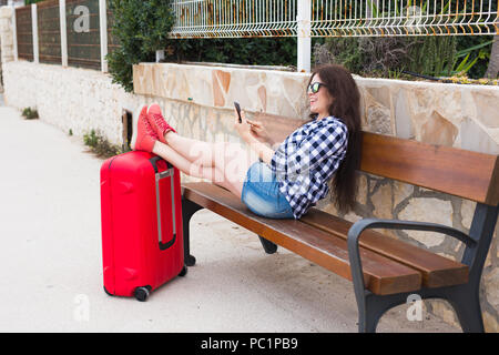 Travel, tourism, technology and people concept - happy woman sit on bench and put her feet on suitcase and typing something on mobile, ready to travel Stock Photo