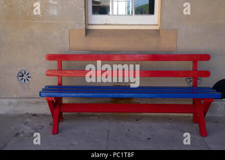 a red and blue wooden bench against cement wall with window in background Stock Photo