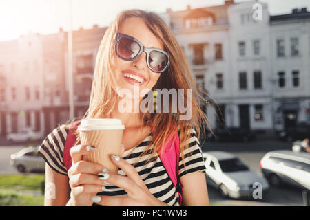 Portrait of closeup teenager long-hair girl in sunglasses drinking coffee and walking on the street with pink backpack Stock Photo