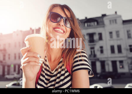 Attractive trendy young woman in sunglasses laughing as she drinking coffee and walking on the street with pink backpack, she having a great time Stock Photo