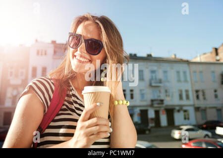 Attractive trendy young woman in sunglasses laughing as she drinking coffee and walking on the street with pink backpack, she having a great time Stock Photo