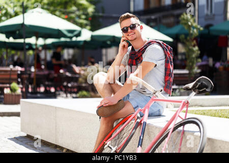 Handsome young hipster man sitting near the bicycle and using smartphone in the sunny street Stock Photo