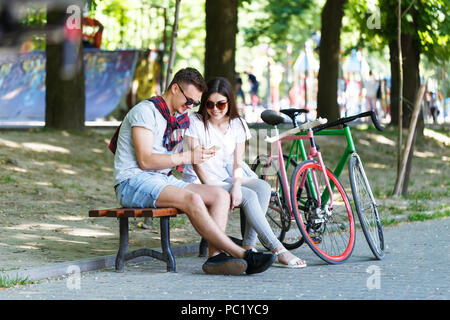 Young couple in love with bicycle sitting on beanch and looking something in the smartphone in the sunny summer park Stock Photo