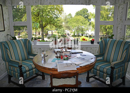 A breakfast nook in front of large windows overlooking the garden with two wing back chairs and circular table cluttered with elements of daily living Stock Photo