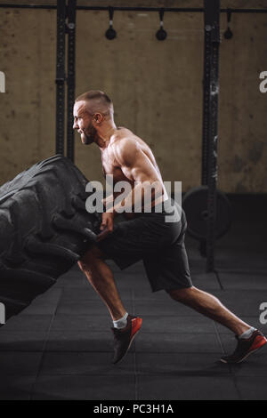 young handsome healthy man with tensed face performing exercise with tractor tire. side view full length photo Stock Photo