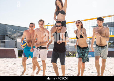 Group of young friends walking on the beach volleyball court in the sunny morning. Two caucasian girls in playful mood and four athletic boys going to