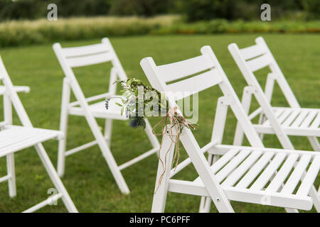 White wedding chairs decorated with fresh flowers on a green grass. Empty wooden chairs for guests on green lawn in the garden prepared for wedding ceremony. Stock Photo