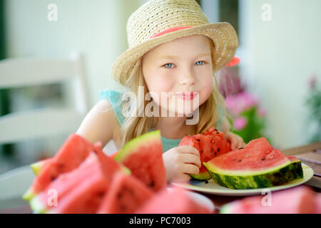 Funny little girl wearing straw hat biting a slice of watermelon outdoors on warm and sunny summer day. Healthy organic food for little kids. Stock Photo