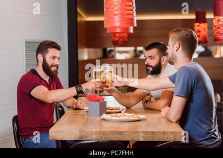 Group of male friends clinking glasses of tasty beer before drinking. Bearded men having appetizing pizza on their table. Three friends having fun and hanging out at pizzeria. Stock Photo