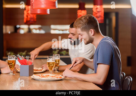 Side view of two male friends eating delicious pizza with help of knife and fork. They also have glasses of tasty beer on wooden table. Interior of modern pizzeria or cafe.   Stock Photo