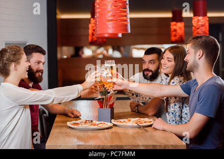 Group of five friends sitting in modern pizzeria or cafe and having fun. Women and men clinking their glasses of tasty beer. They also have two appetizing pizzas on wooden table. Stock Photo