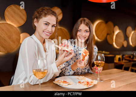 Two beautiful young women smiling, looking at camera and posing.Gorgeous female friends eating delicious pizza  and spending time in pizzeria. Glasses of white wine placed on wooden table. Stock Photo
