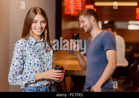 Front view of gorgeous young woman posing, looking at camera and smiling. Smiling man taking photo of his friend using phone. Positive girl holding black paper cup of coffee. Cafe interior.