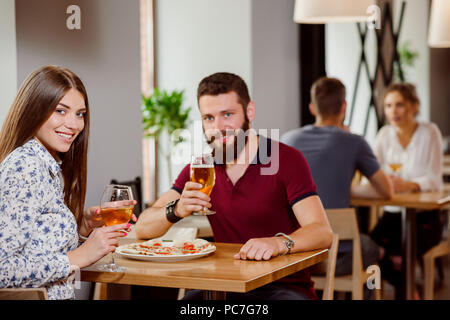 Beautiful couple sitting in pizzeria and posing with glasses of white wine and beer. Gorgeous woman and bearded man looking at camera and smiling. People talking in restaurant on background. Stock Photo