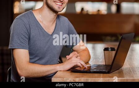 Positive man sitting in cafe, drinking coffee and working with notebook. Programmer holding pen, smiling and typing on computer.  Concept of workplace in cafe and programming. Stock Photo
