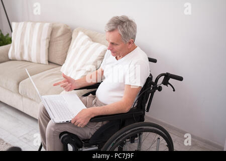 Man in wheelchair working with computer. Senior disabled man sitting in wheelchair in his living room and doing some work on his white laptop. Stock Photo